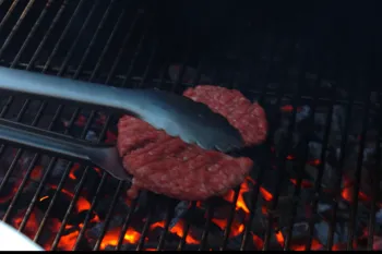 Tongs laying a raw beef patty onto a charcoal grill over glowing coals
