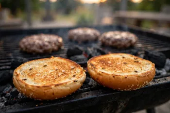 Bun halves toasting on the grill with patties behind them