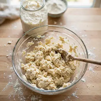 Shaggy just-mixed dough in a glass bowl with a wooden spoon, the starter jar behind it